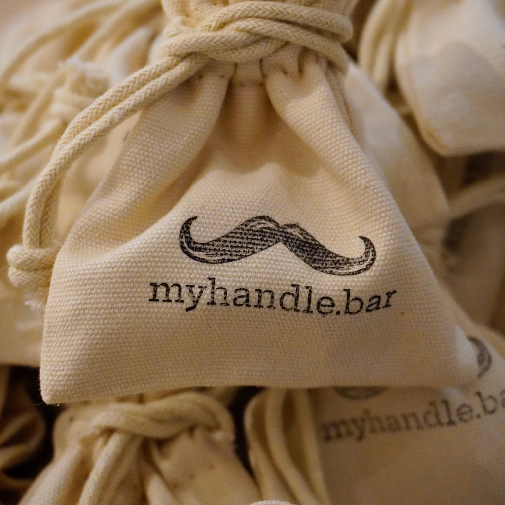 Beige linen drawstring bags with 'myhandle.bar' logo on a beige background. Travel bags for small moustache grooming products such as Training Wheels, combs, wax, oil, scissors.